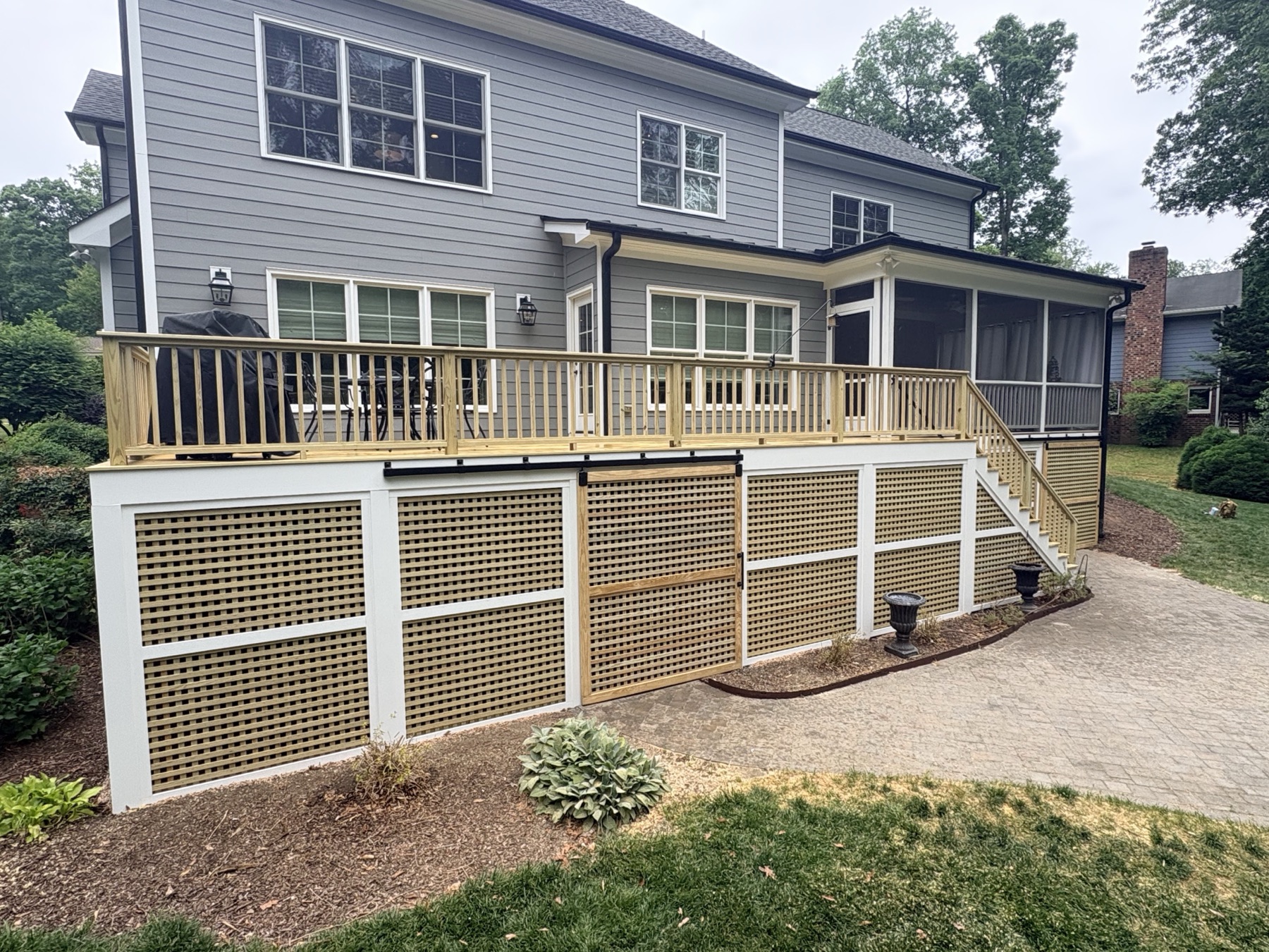 A raised deck across the back of a two-story home, finished with a crisp white-framed lattice skirt over the paver drive.
