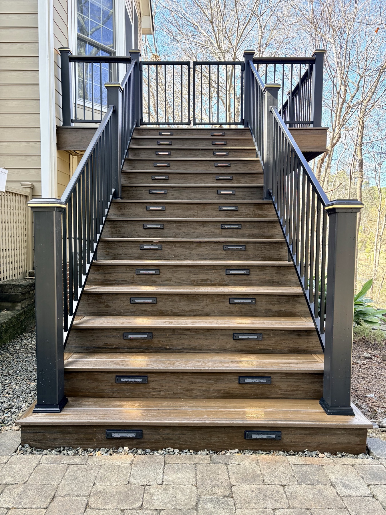 A wide staircase in composite decking with lit treads and dark metal balusters, running down to a paver landing.