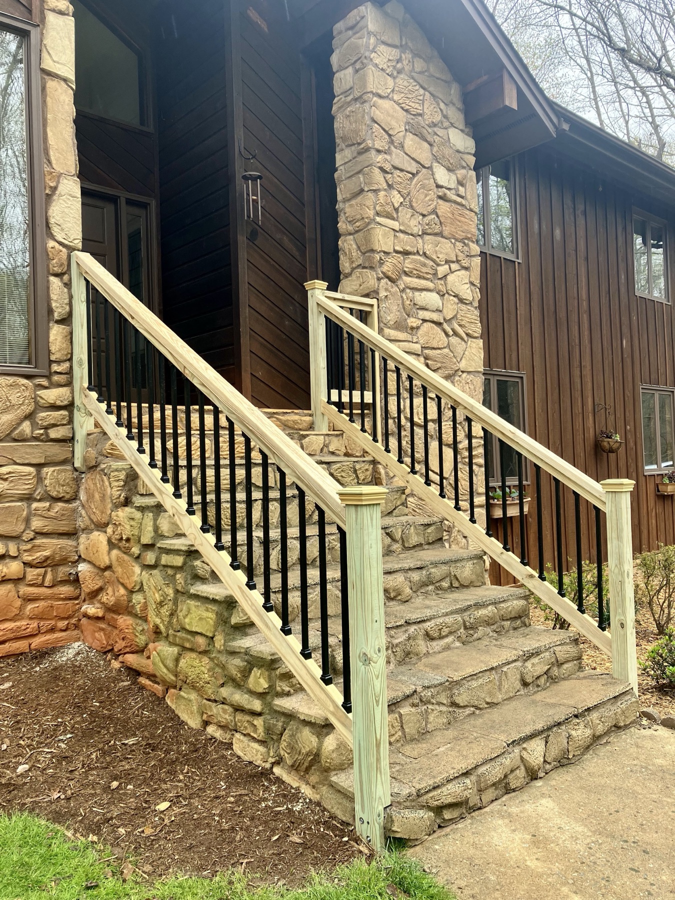 A fresh wood staircase with metal balusters flanked by stone, tying a rustic home cleanly to the yard.